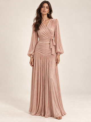 A woman modeling a floor-length dusty pink pleated wrap dress with long balloon sleeves and a tied waist, photographed against a plain warm beige studio background.