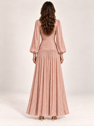 A woman viewed from behind wearing a long dusty pink pleated maxi dress with voluminous balloon sleeves and a gathered bodice, paired with tan high heels against a light neutral studio background.
