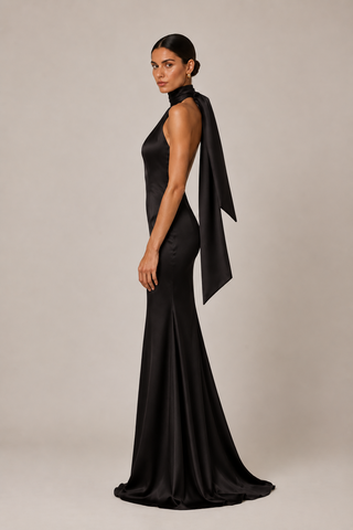 A woman modeling a floor-length black satin gown with a high halter-style neckline and a dramatic draped fabric detail over one shoulder, photographed against a plain light gray studio background.