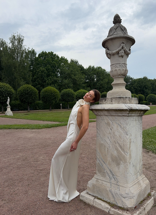 A woman in a sleeveless cream satin gown leans against a large white marble garden urn pedestal in a formal park with green lawns and trees under a cloudy sky.