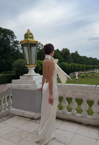 A woman wearing a white satin backless gown with a large bow at the back stands on a stone terrace in a park, seen from behind. A decorative lamp post is beside her, with a manicured garden and trees in the background.