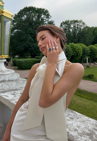 A woman in a cream satin dress with a large bow collar poses outdoors, wearing multiple silver rings on her fingers. The background features a stone wall, a green garden, and a classical lamp post.