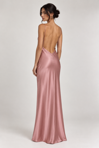 A woman modeling a floor-length satin gown from the back, showing a low open back and thin straps, in a dusty rose pink color against a light gray studio background.