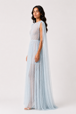 A woman modeling a light blue sheer maxi dress with a high neckline, cap sleeves, and a flowing tulle skirt against a plain light gray studio background.