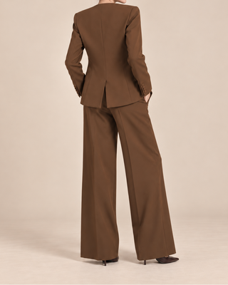 A woman stands with her back to the camera wearing a tailored brown suit with a structured blazer and wide-leg trousers, photographed against a plain warm beige studio background.