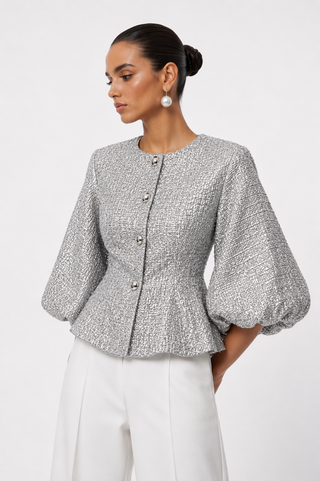 A woman wearing a textured silver-gray cropped jacket with puff sleeves and a button front, paired with white trousers, photographed against a light gray studio background.