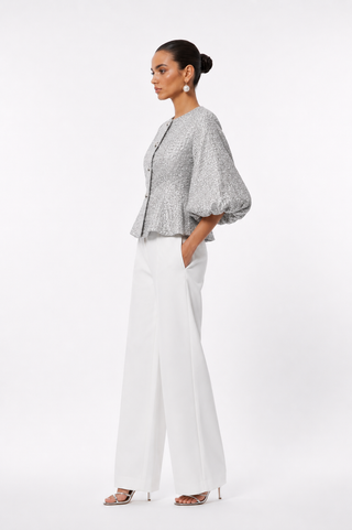 A woman modeling a light gray patterned short-sleeve blouse with dramatic puffed sleeves and white wide-leg trousers, standing against a plain white studio background.