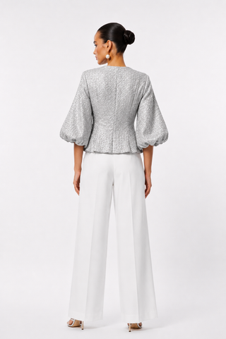 A woman shown from the back wearing a cropped silver textured jacket with voluminous puff sleeves and white wide-leg trousers, standing against a light gray studio background.