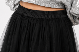 Close-up of a black pleated tulle skirt with an elastic waistband, shown on a model wearing a silver textured top against a light gray background.