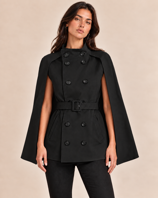 A woman modeling a black double-breasted cape jacket with a matching waist belt, photographed against a plain beige studio background.