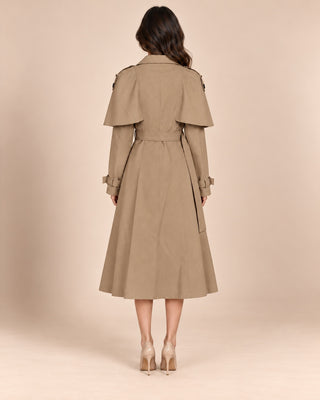 A woman viewed from the back wearing a mid-length beige trench coat with wide shoulder panels and a self-tie waist belt, paired with nude high heels against a plain warm beige studio background.