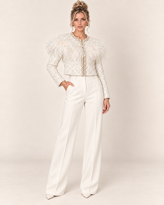 A woman models a white embellished jacket with dramatic feathered shoulders and matching white wide-leg trousers against a warm beige studio background.