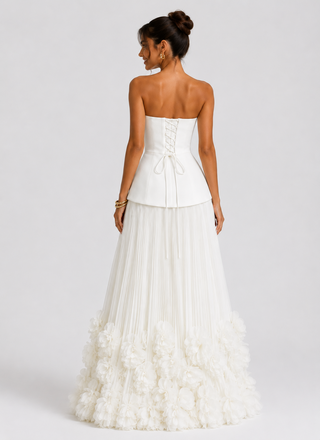 A woman modeling a strapless white wedding gown from the back, showing a structured corset-style bodice with lace-up detailing and a voluminous layered tulle skirt with ruffled floral texture at the bottom, against a light gray studio background.