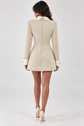 A woman viewed from the back wearing a beige tweed mini dress with cream-colored collar and cuffs, paired with cream high heels, against a light gray studio background.