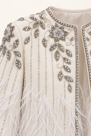 Close-up of a cream-colored embellished garment featuring intricate silver beadwork, sequins, and white feathers along the front.