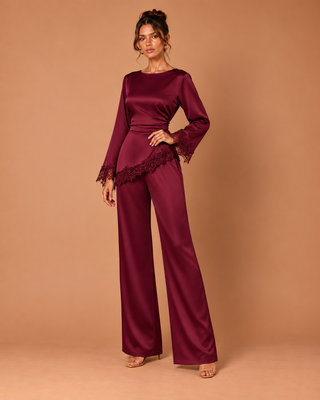A woman models a deep burgundy satin jumpsuit with long bell sleeves, lace trim at the cuffs and hem, and wide-leg pants, set against a solid warm tan studio background.
