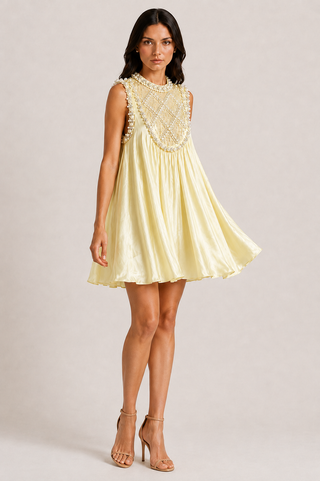 A woman modeling a pale yellow sleeveless mini dress with a beaded neckline and a flared skirt, paired with nude high-heeled sandals, against a light gray studio background.