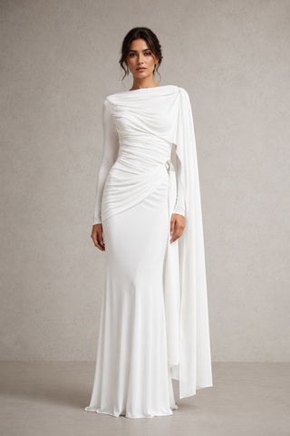 A woman modeling a floor-length white draped gown with long sleeves, a fitted silhouette, and a flowing cape-like overlay, photographed against a neutral textured studio backdrop.