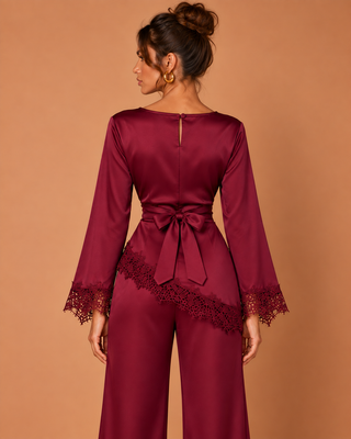 A woman shown from the back wearing a satin-like burgundy jumpsuit with long sleeves, lace-trimmed cuffs, a matching waist tie, and wide legs, against a warm tan studio background.