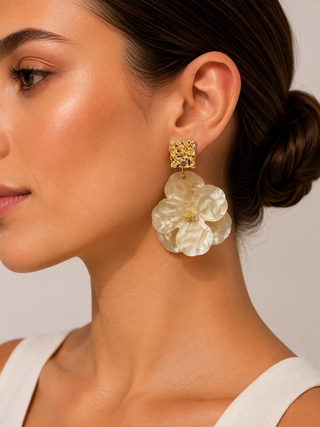 A close-up of a woman wearing a large statement earring with a textured gold square stud and a layered off-white flower drop, shown against a neutral light gray background.