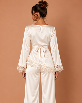 A woman shown from the back wearing a cream satin jumpsuit with lace trim on the cuffs and hem, tied at the waist. She is standing against a solid warm tan background.