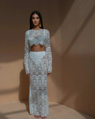 A woman wearing a white crochet floral two-piece beach dress featuring a long-sleeve crop top and a semi-sheer maxi skirt, styled in a bohemian Ibiza beach club aesthetic against a warm-toned background. Vienna Fashion