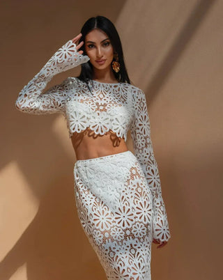 A woman wearing a white crochet floral two-piece beach dress featuring a long-sleeve crop top and a semi-sheer maxi skirt, styled in a bohemian Ibiza beach club aesthetic against a warm-toned background. Vienna Fashion