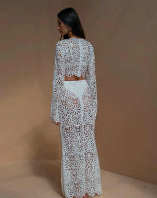 A woman wearing a white crochet floral two-piece beach dress featuring a long-sleeve crop top and a semi-sheer maxi skirt, styled in a bohemian Ibiza beach club aesthetic against a warm-toned background. Vienna Fashion