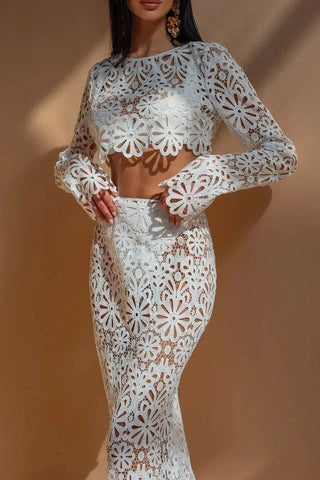 A woman wearing a white crochet floral two-piece beach dress featuring a long-sleeve crop top and a semi-sheer maxi skirt, styled in a bohemian Ibiza beach club aesthetic against a warm-toned background. Vienna Fashion