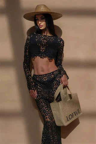 A woman wearing a black crochet floral two-piece beach dress featuring a long-sleeve crop top and a semi-sheer maxi skirt, styled in a bohemian Ibiza beach club aesthetic against a warm-toned background.