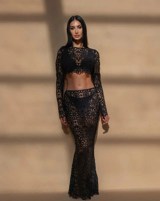 A woman wearing a black crochet floral two-piece beach dress featuring a long-sleeve crop top and a semi-sheer maxi skirt, styled in a bohemian Ibiza beach club aesthetic against a warm-toned background.