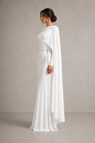A woman modeling a floor-length white draped gown with long sleeves and a flowing cape-like overlay, photographed in profile against a neutral textured studio backdrop.