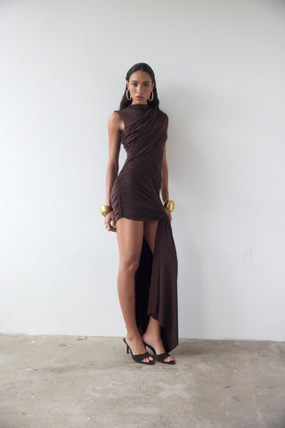 Woman wearing a brown dress with a slit standing against a white wall.
