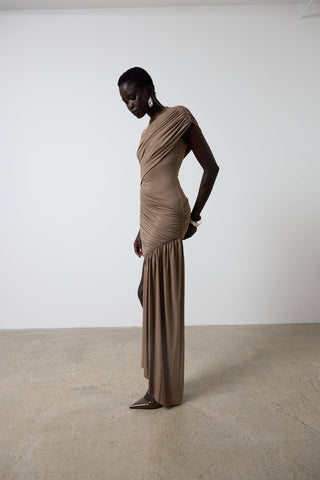 Woman wearing a brown draped dress standing against a white wall.