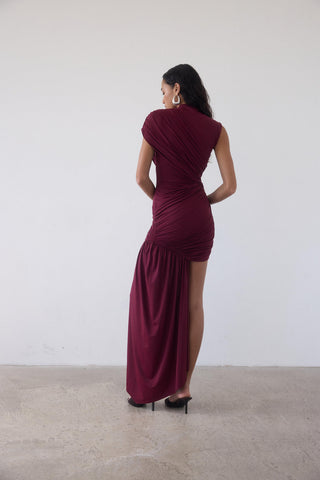 Woman wearing a burgundy evening gown with a slit, standing against a plain white background.