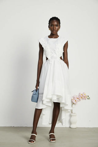 Model wearing a sleeveless white dress with a round neck and semi-sheer construction, featuring lace trim and pearl embellishments. The dress has an asymmetric hem, flounce detailing, and a backless design, perfect for summer vacations and elegant occasions.