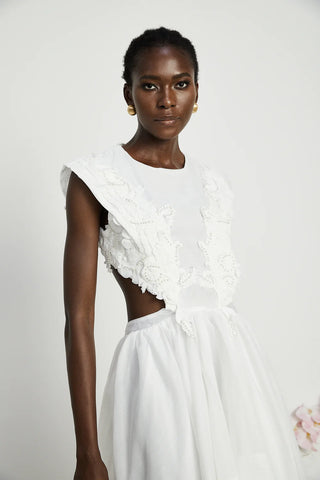 Model wearing a sleeveless white dress with a round neck and semi-sheer construction, featuring lace trim and pearl embellishments. The dress has an asymmetric hem, flounce detailing, and a backless design, perfect for summer vacations and elegant occasions.