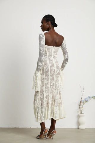 Elegant woman wearing a white bohemian lace maxi dress with long bell sleeves, intricate floral lace pattern, and a sweetheart neckline, standing gracefully in a minimalistic setting with a small vase of flowers. - Cielie Vienna