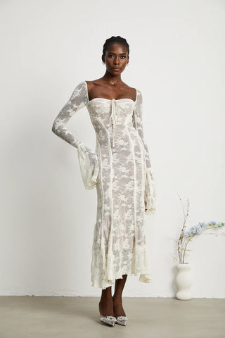 Elegant woman wearing a white bohemian lace maxi dress with long bell sleeves, intricate floral lace pattern, and a sweetheart neckline, standing gracefully in a minimalistic setting with a small vase of flowers. - Cielie Vienna