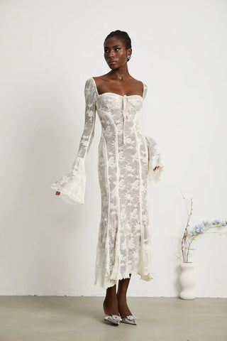 Elegant woman wearing a white bohemian lace maxi dress with long bell sleeves, intricate floral lace pattern, and a sweetheart neckline, standing gracefully in a minimalistic setting with a small vase of flowers. - Cielie Vienna