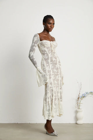 Elegant woman wearing a white bohemian lace maxi dress with long bell sleeves, intricate floral lace pattern, and a sweetheart neckline, standing gracefully in a minimalistic setting with a small vase of flowers. - Cielie Vienna