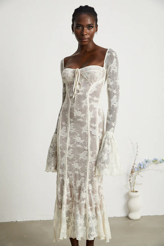 Elegant woman wearing a white bohemian lace maxi dress with long bell sleeves, intricate floral lace pattern, and a sweetheart neckline, standing gracefully in a minimalistic setting with a small vase of flowers. - Cielie Vienna