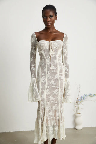 Elegant woman wearing a white bohemian lace maxi dress with long bell sleeves, intricate floral lace pattern, and a sweetheart neckline, standing gracefully in a minimalistic setting with a small vase of flowers. - Cielie Vienna