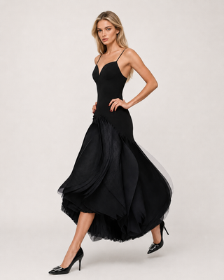 A woman modeling a black spaghetti-strap midi dress with a structured bodice and a layered, flowing tulle skirt, paired with black pointed-toe heels, against a light gray studio background.