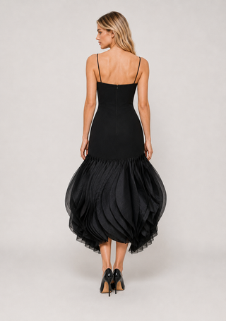 A woman modeling a black spaghetti-strap midi dress from the back, featuring a structured bodice and a voluminous ruffled tulle skirt, set against a plain light gray studio background.